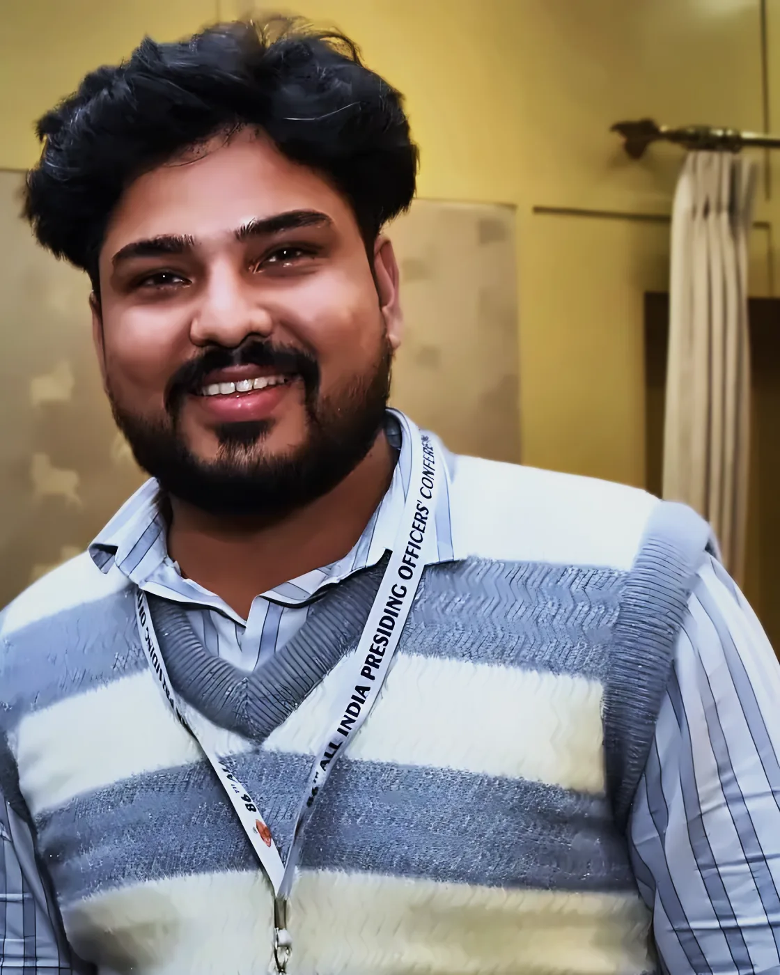 Portrait of Prem Mishra, smiling, wearing a striped sweater vest and a conference delegate lanyard